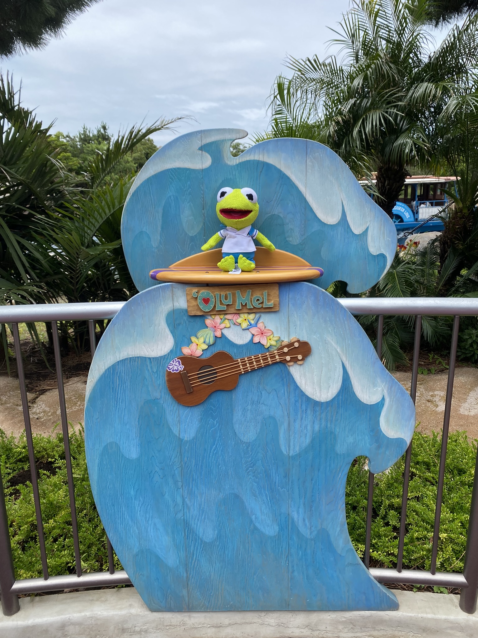 Kermit sitting on a fake, miniature surfboard with a fake wave behind him.