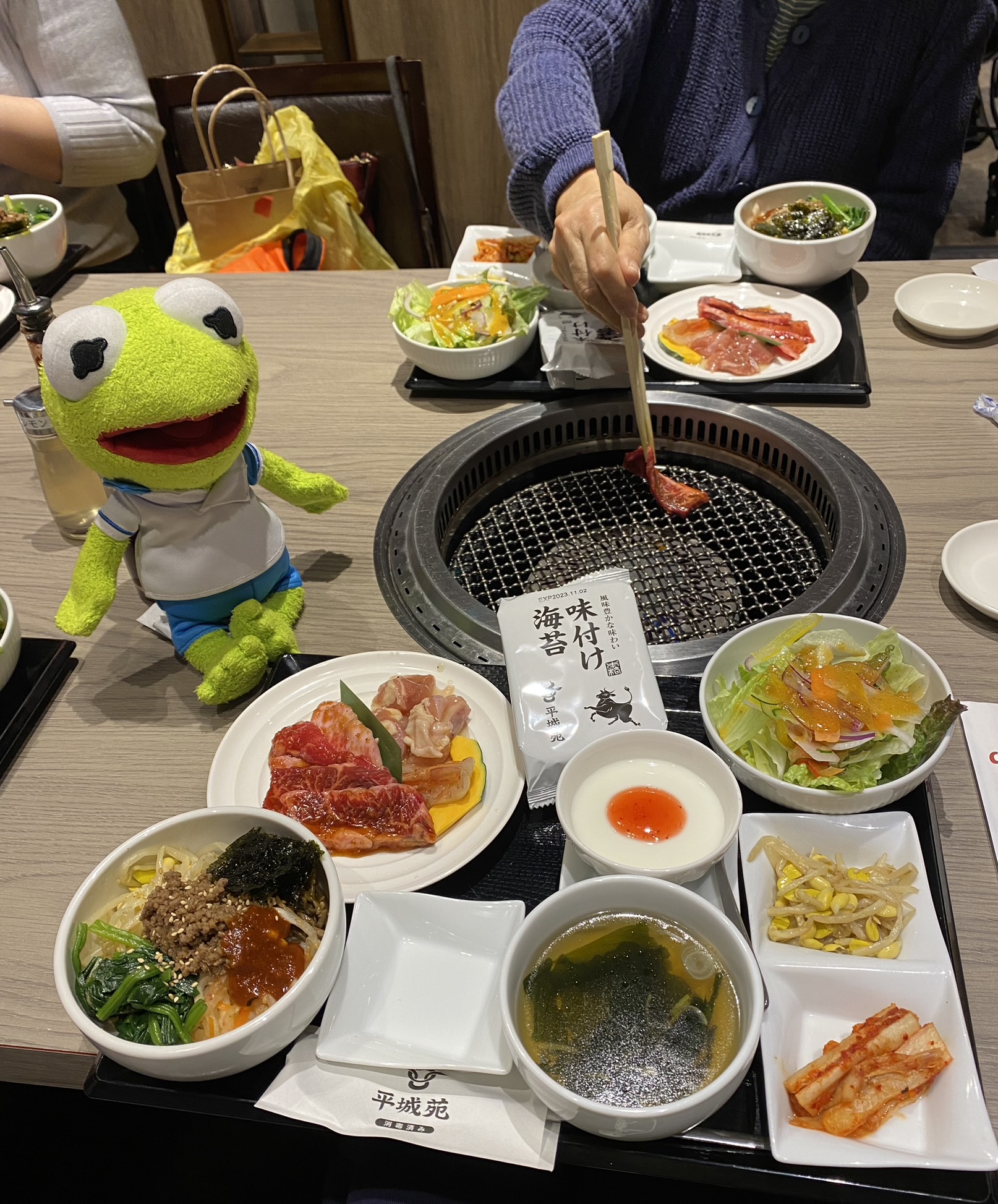 Kermit standing by a tray of raw beef and other side dishes. Behind Kermit is a grill where someone is placing raw meat.