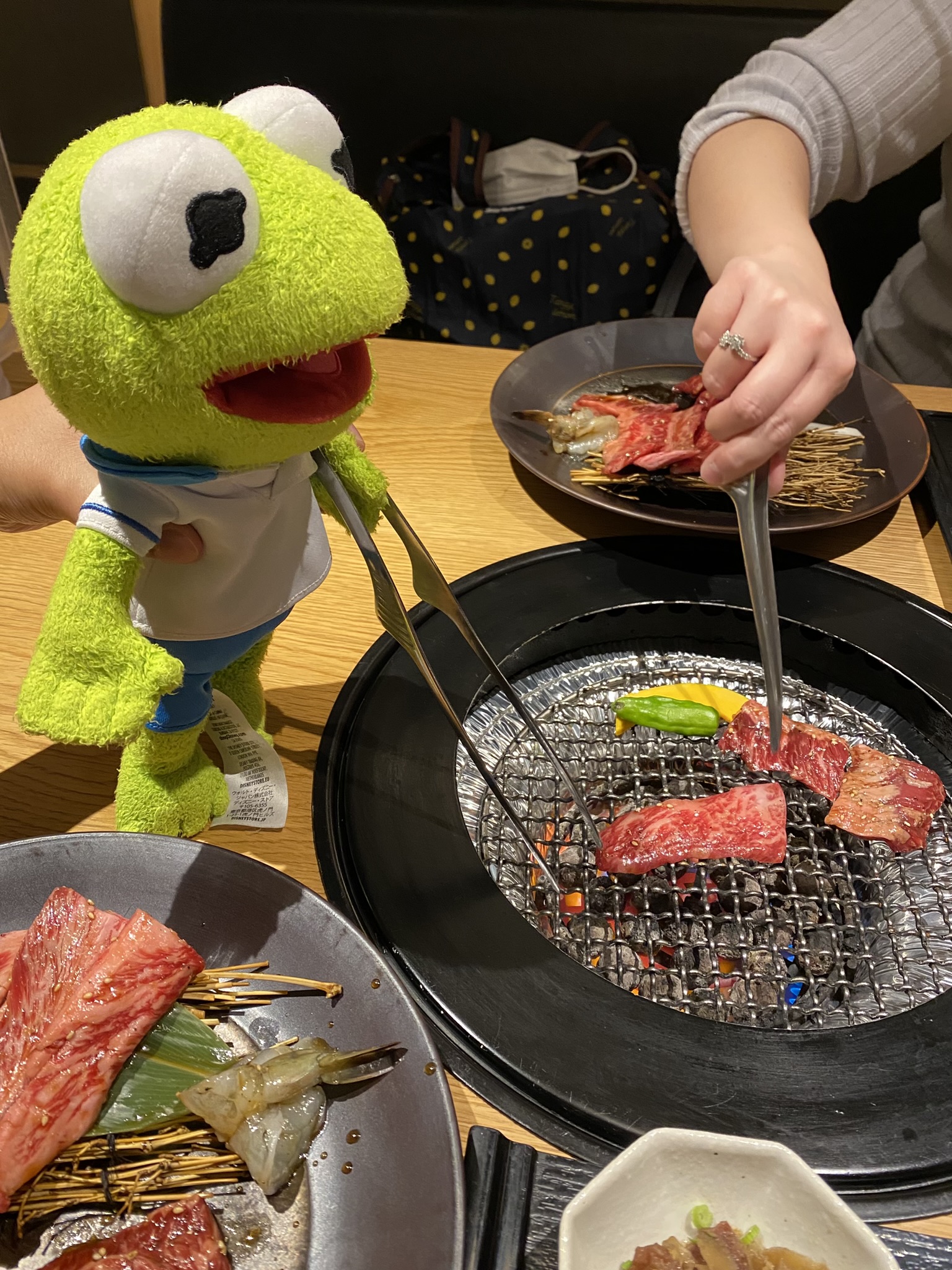 Kermit standing next to platter of raw meat and seafood while grilling the meat with tongues.