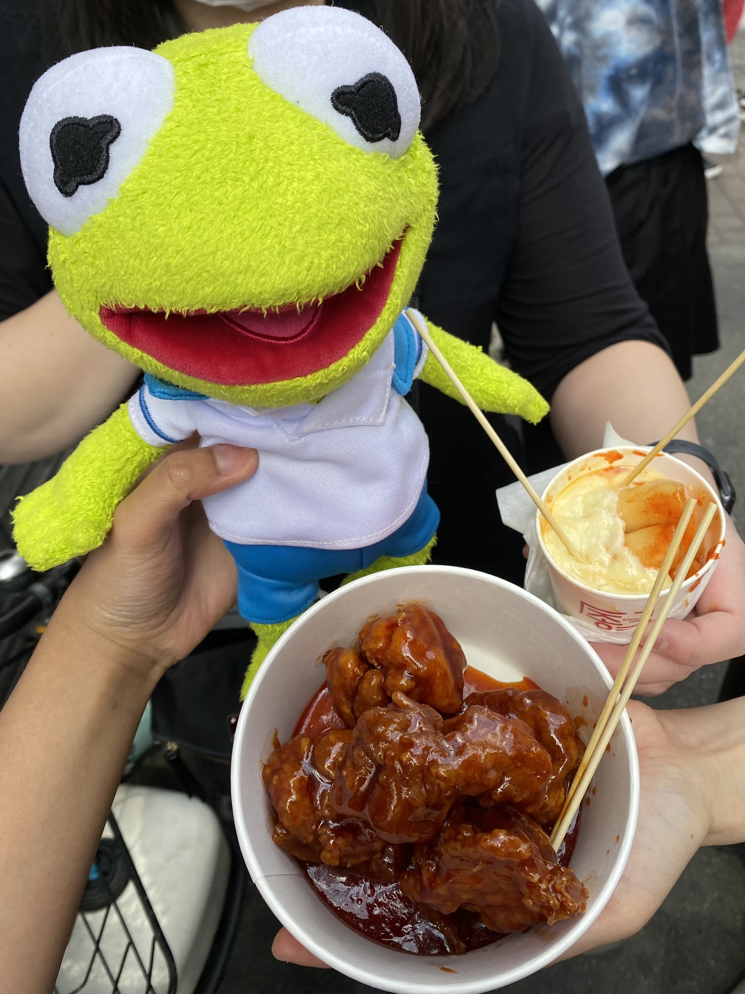 Kermit posing for a picture next to tteokkbokki covered in melted cheese and crispy Korean fried chicken.