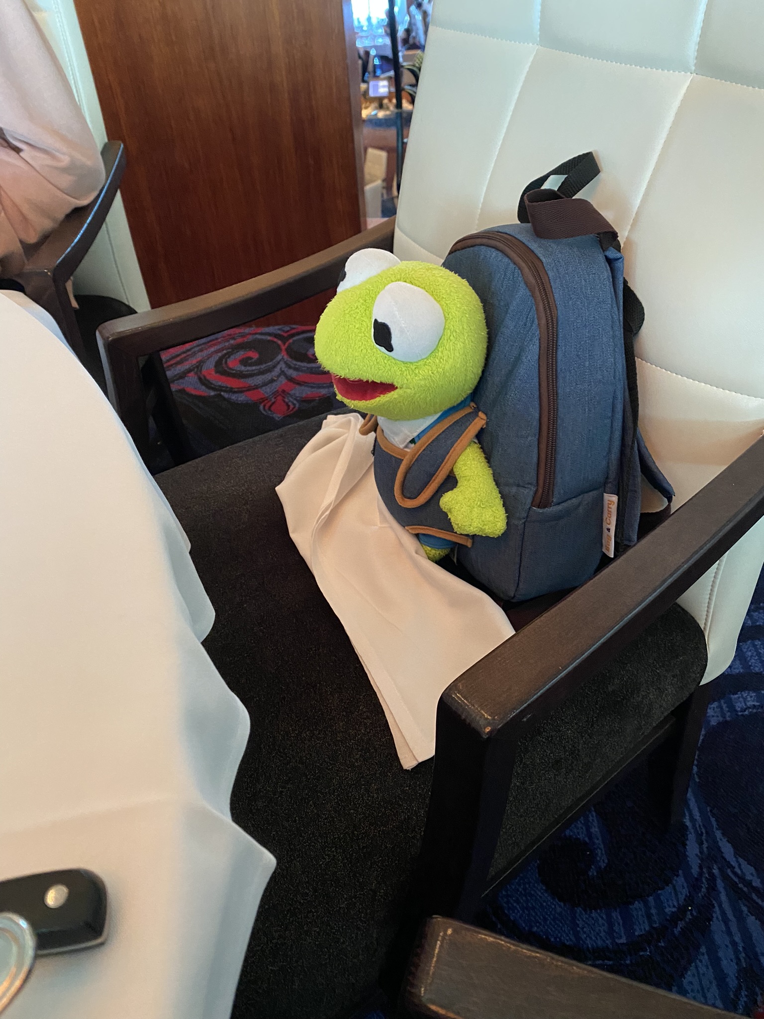 Kermit sitting in a chair that is way too large for him. He is at the dinner table in a restaurant with a napkin in his lap.