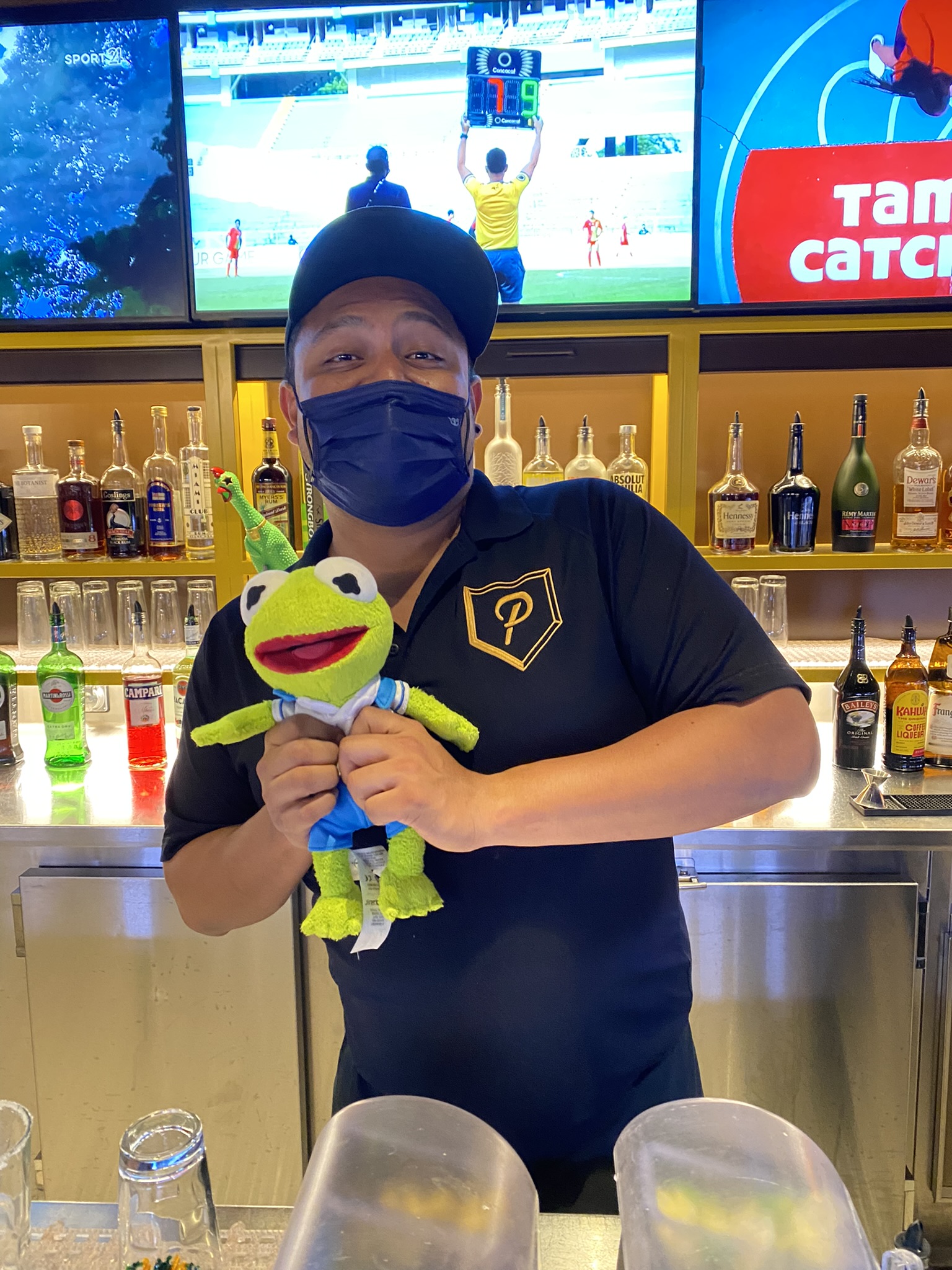 Kermit being held by a bartender wearing a black polo shirt behind the bar.