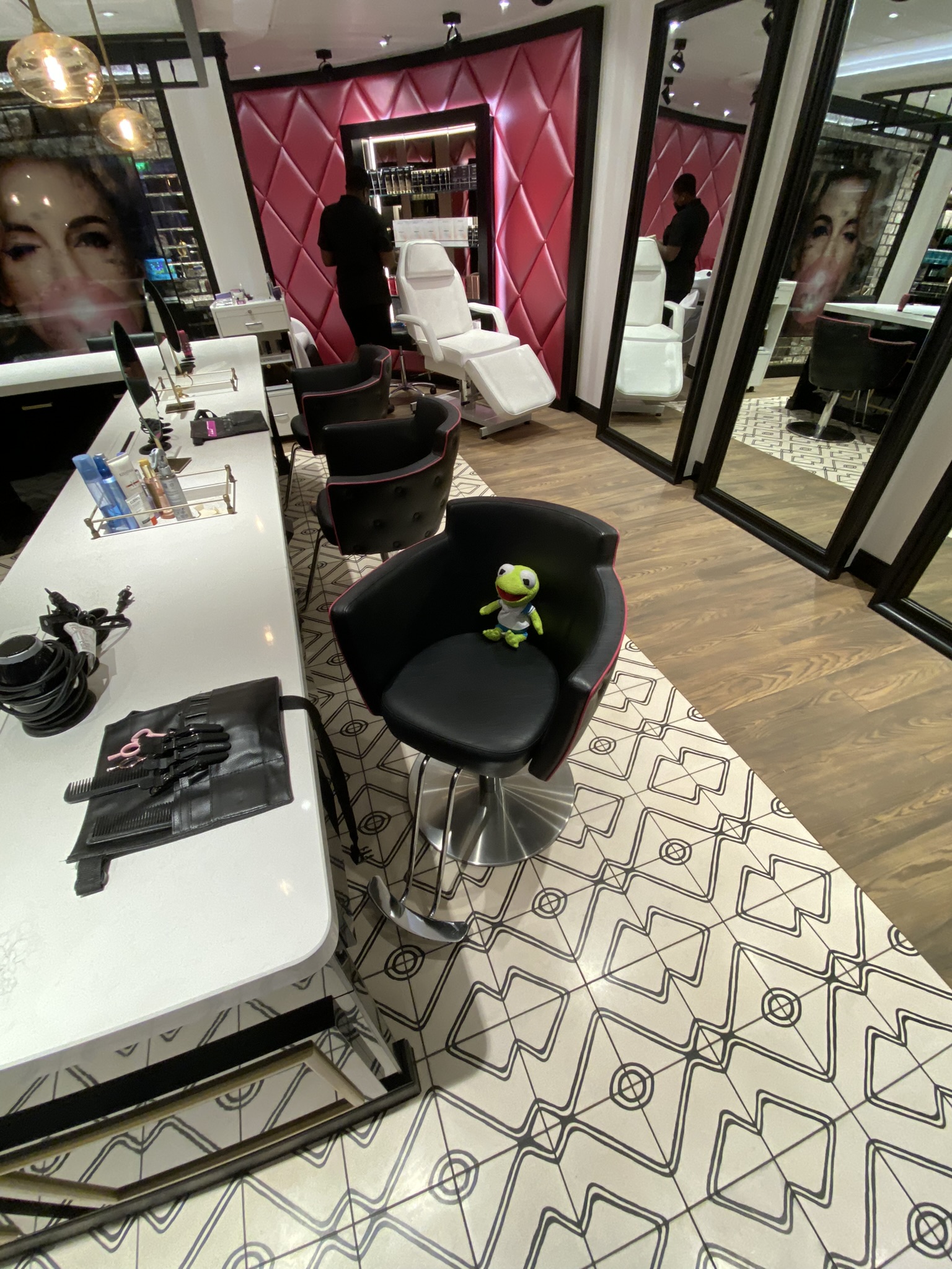 Kermit sitting in a salon chair in the middle of a beauty salon. He is sitting before a white counter, the chair is black leather, and there are many ceiling to floor mirrors.