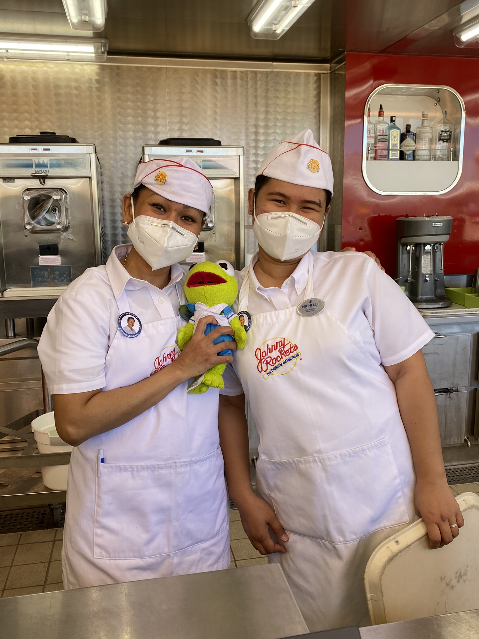 Kermit being held by two women wearing Johnny Rocket's uniforms at Johnny Rockets.